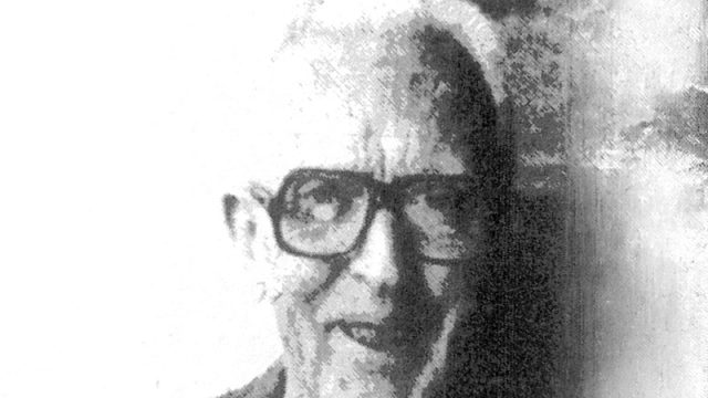 A black and white, grainy photo of an older man with short white hair, wearing large dark glasses, a suit, and a tie. The left side of the image is bright, and the right side appears darker and blurred.
