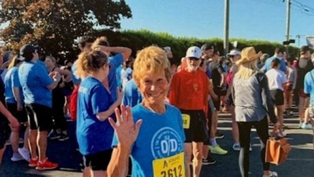 An older woman wearing a blue shirt and race bib number 2612 smiles and waves at the camera, surrounded by a crowd of other runners gathered at an outdoor event.