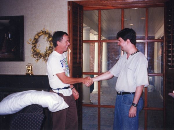 Two men are smiling and shaking hands indoors near wooden doors. One man wears a white t-shirt and brown pants, the other a light gray shirt and jeans. There’s a pillow on a chair and a wreath on the wall behind them.