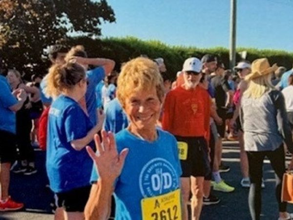 An older woman wearing a blue shirt and race bib number 2612 smiles and waves at the camera, surrounded by a crowd of other runners gathered at an outdoor event.