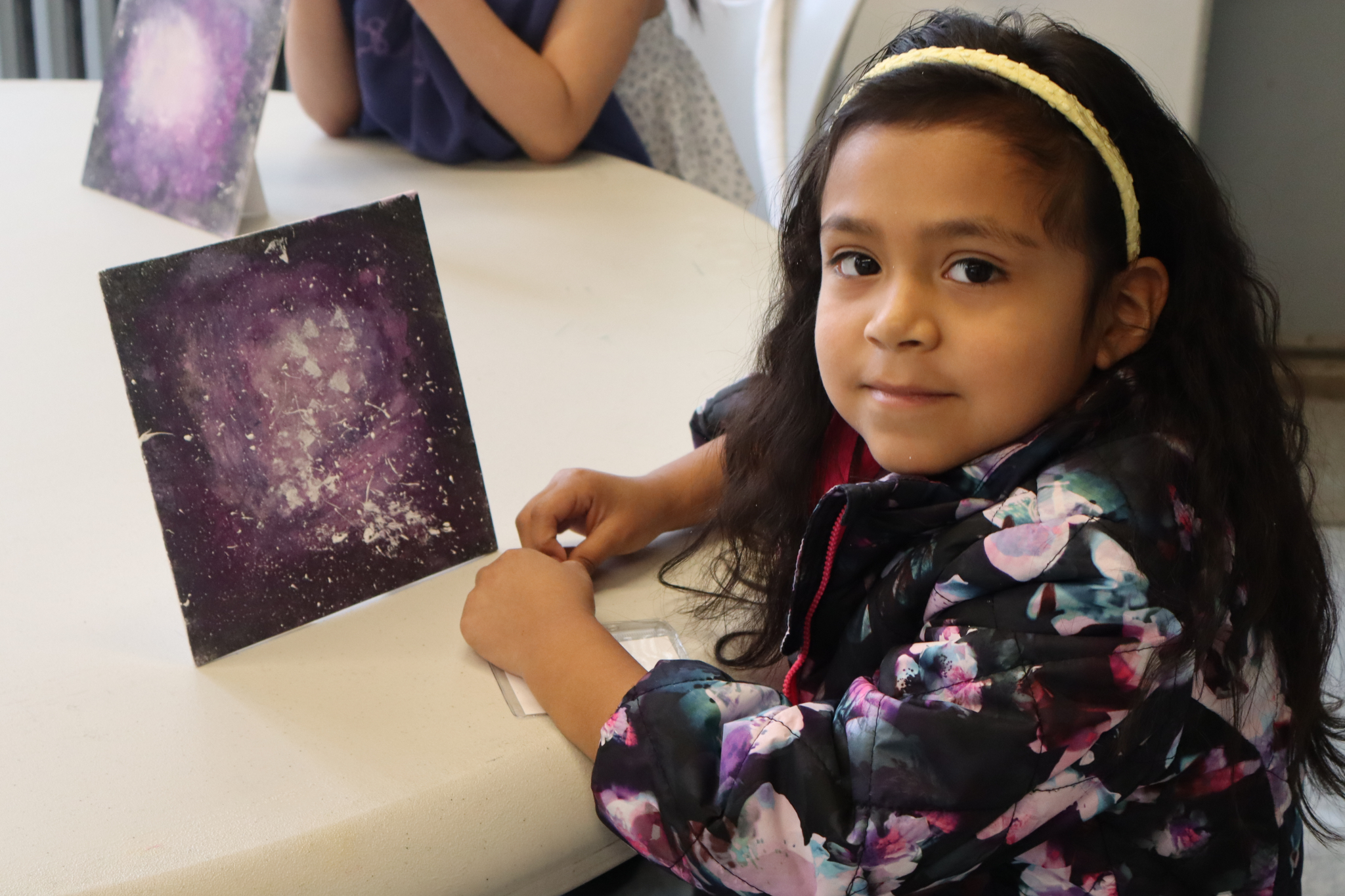 A young girl wearing a floral jacket and yellow headband sits at a table, smiling, with a painting of a purple galaxy propped up in front of her. Another child with a similar artwork is visible in the background.