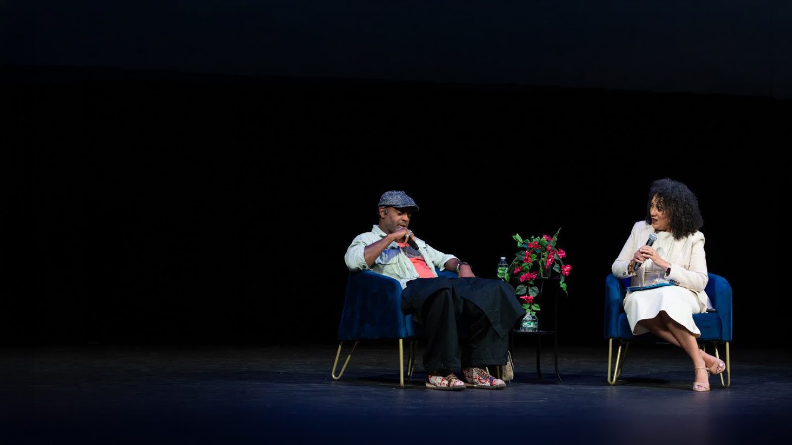 Two people sit on stage in blue chairs, engaged in conversation. A small table with a bouquet of flowers sits between them. The background is dark, highlighting the speakers under stage lighting.