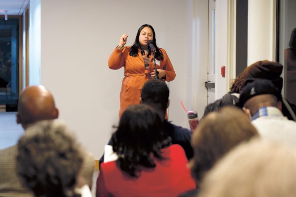 A woman in an orange dress stands and speaks into a microphone in front of a seated audience in a brightly lit room. She gestures with her hand while people listen attentively.