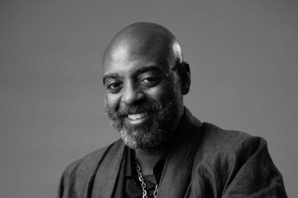 A middle-aged man with a bald head and a gray beard smiles warmly at the camera. He is wearing a jacket and a necklace, and the photo is in black and white with a plain background.