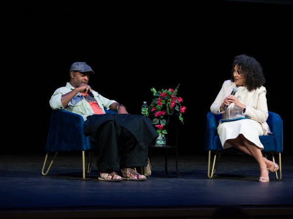 Two people sit on stage in blue chairs, engaged in conversation. A small table with flowers and water bottles is between them. The background is dark, and both are holding microphones.