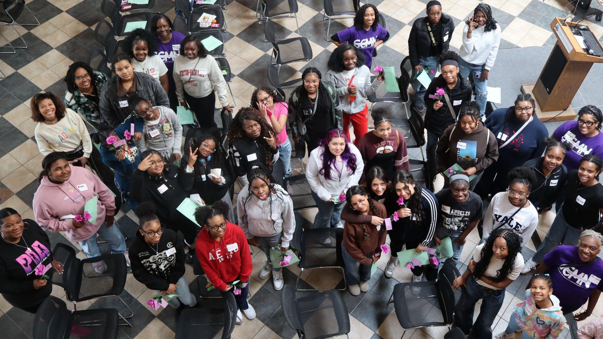 A group of young women stand together in a tiled room, smiling and looking up at the camera. Many hold pink flowers and wear name tags, and empty chairs are scattered around them.
