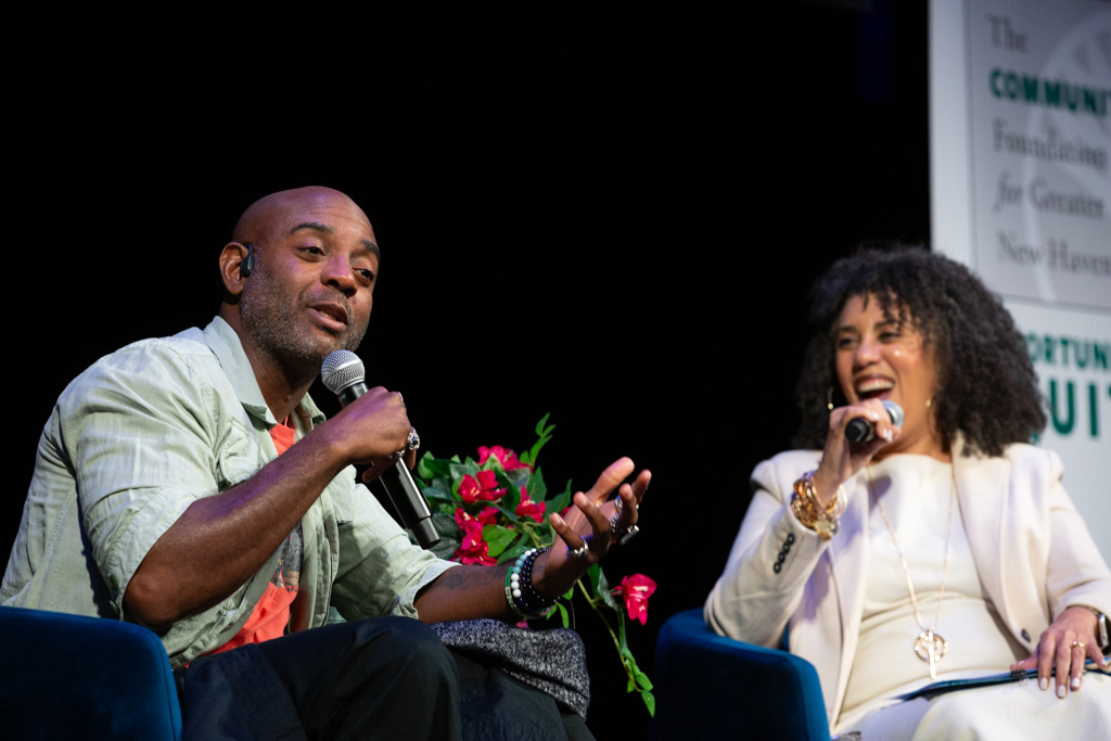 A man and a woman sit on stage, holding microphones and engaging in a lively conversation. The man gestures with his hand while speaking, and the woman smiles broadly. A bouquet of red roses sits between them.