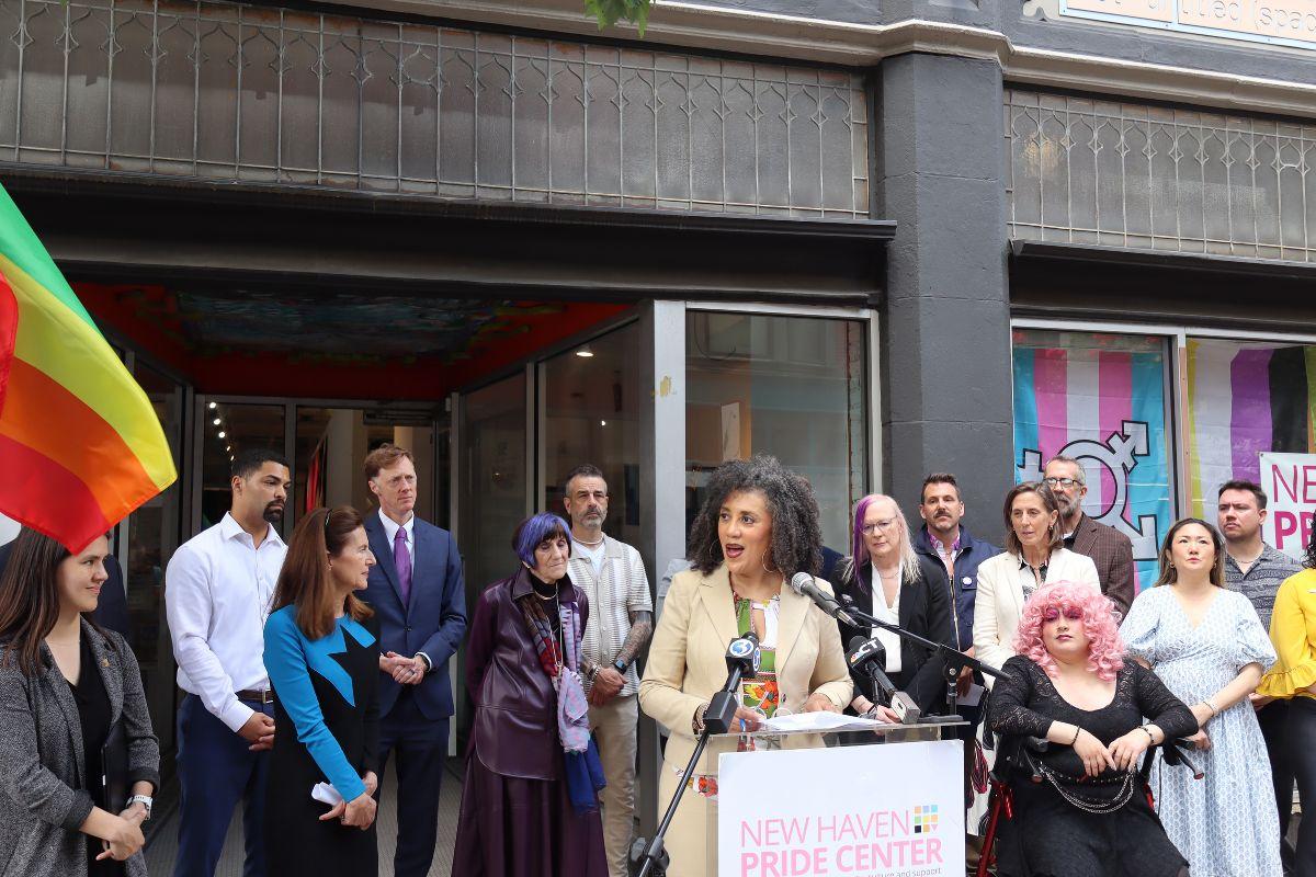 A group of people stand outside the New Haven Pride Center. A speaker addresses the crowd at a podium, with pride flags, microphones, and others listening and standing nearby. The building displays LGBTQ+ symbols and signage.