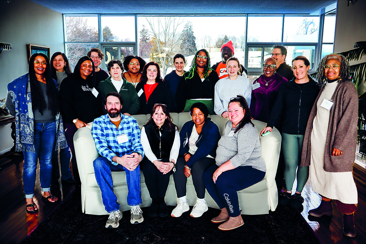 A group of nineteen adults pose and smile together indoors, with some seated on a white couch and others standing behind it. Large windows in the background let in daylight and show trees outside.