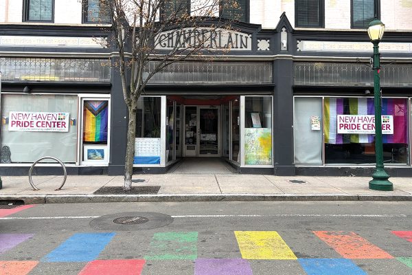 Street view of the New Haven Pride Center, with rainbow pride flags and signs in the windows. A tree stands on the sidewalk, and a rainbow crosswalk is painted on the street in front of the building.