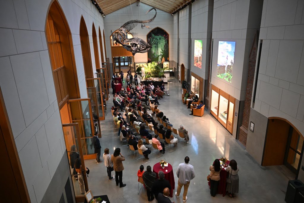 A group of people sit and stand in a large museum hall with dinosaur skeletons overhead, listening to a speaker at the front. The hall has tall windows, arched doorways, and display cases along the walls.