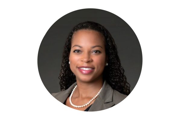 Professional headshot of a woman with long curly hair, wearing a gray blazer, white blouse, pearl necklace, and pearl earrings, smiling against a dark background. The photo is framed in an oval shape.
