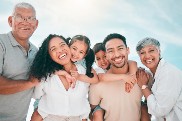 Three generations of a smiling family pose together outdoors under a bright sky. Two children are piggybacked by adults, surrounded by two elders, all looking happy and close.