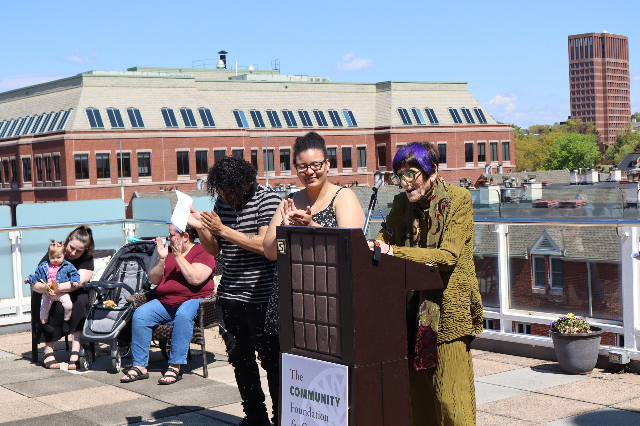 Three people stand at a podium on a sunny rooftop, one speaking into a microphone. In the background, a few seated individuals, a baby in a stroller, and brick buildings are visible under a clear blue sky.