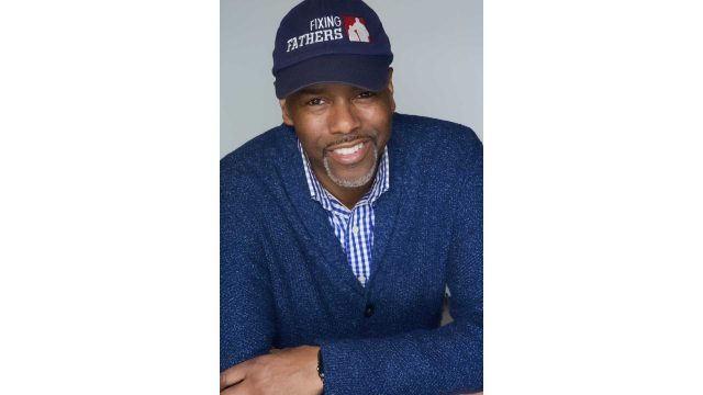 A man in a blue sweater and checkered shirt, wearing a navy Fixing Fathers cap, smiles while sitting with his arms crossed on a table.
