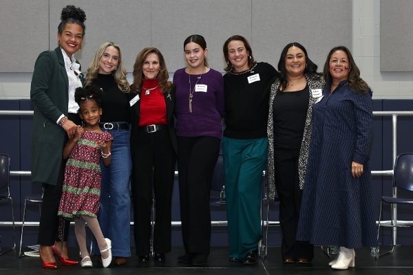 Seven women and a young girl stand together indoors, smiling at the camera. The group is casually dressed, with name tags visible, suggesting a friendly event or gathering.