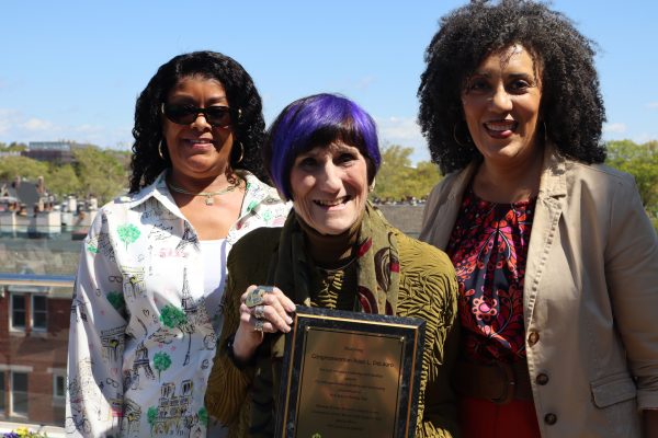 Three women stand together outdoors in sunlight, smiling at the camera. The woman in the center with short purple hair holds a plaque, while the two women beside her wear glasses and patterned jackets. Buildings and trees are visible behind them.