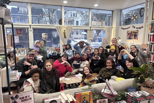 A diverse group of people, including children and adults, gather inside a cozy bookstore. They smile, make heart shapes with their hands, and sit among bookshelves and tables covered with books, creating a warm, welcoming atmosphere.