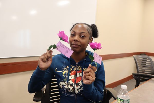 A woman wearing a blue hoodie sits at a table, holding two artificial pink roses with tags. She is smiling slightly, and there is a water bottle on the table in front of her. A whiteboard is visible in the background.