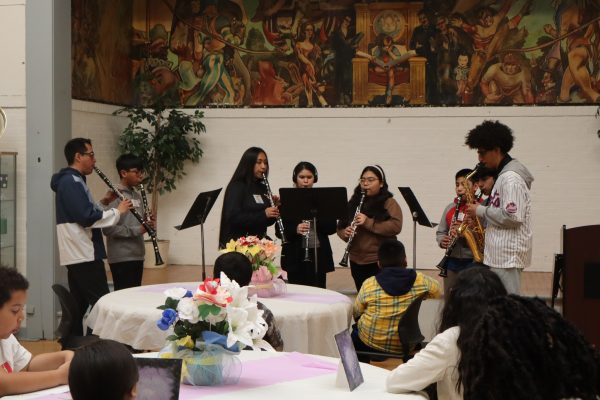 A group of students play wind instruments in front of an audience seated at decorated round tables. A mural is visible on the wall behind them. Some audience members watch attentively.