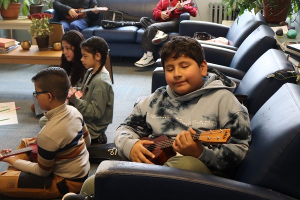 A boy sits on a chair playing a ukulele with his eyes closed, surrounded by other children and an adult also holding ukuleles in a cozy room with plants and books.