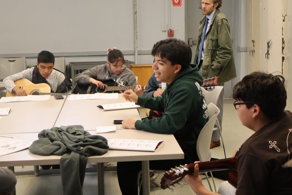 Four students sit around a table playing guitars and reading sheet music, while an adult stands in the background observing them. The students appear engaged and are in a classroom setting.