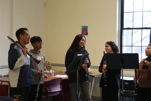 Five students stand indoors holding clarinets, talking and smiling. They are gathered around music stands near a window, with chairs and tables in the background. One student is reading from sheet music.