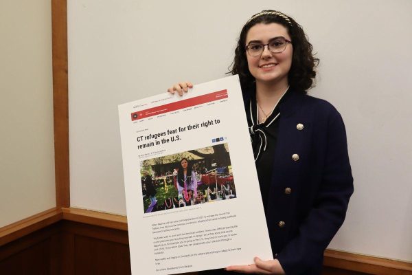 A woman with glasses and dark hair smiles while holding a large poster featuring a news article titled CT refugees fear for their right to remain in the U.S. with a photo and text.