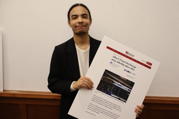 A person with long dark hair tied back, wearing a black blazer and light shirt, stands in front of a whiteboard holding a large printout of an article about city transit, featuring a bus photo and text.