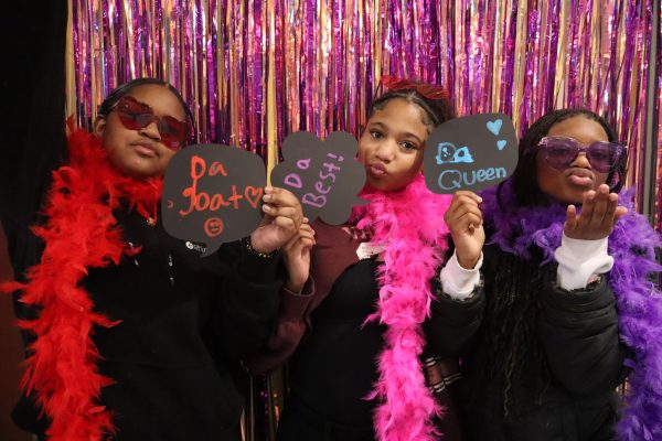 Three girls pose playfully in front of pink and orange streamers, wearing feather boas and sunglasses. Each holds a chalkboard sign reading “Da Goat,” “Da Best!,” and “Da Queen.”.