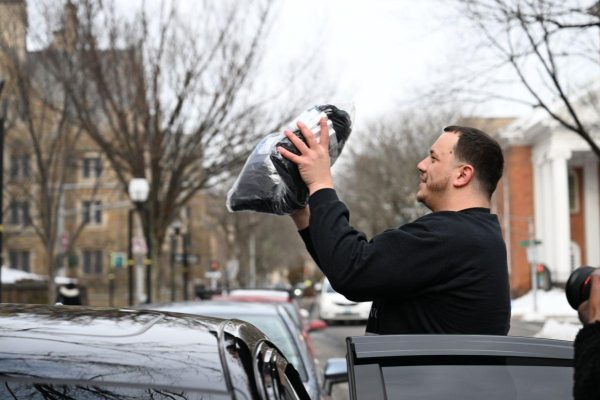 A man stands beside a car holding up a black bag with both hands on a snowy street lined with bare trees and historic buildings.