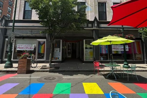 A street scene in front of the New Haven Pride Center, featuring rainbow-colored crosswalk stripes, outdoor tables with bright umbrellas, and a tree providing shade on a sunny day.