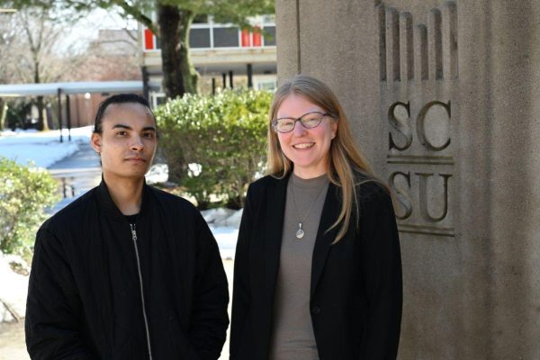 Two people stand outside next to a stone sign with the letters SCSU engraved on it. Trees and a building are visible in the background on a sunny day.