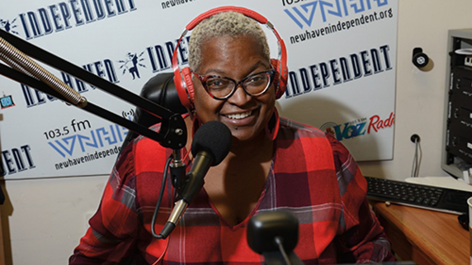 A smiling person with short blond hair and red headphones speaks into a microphone in a radio studio, wearing a red plaid shirt. The background features logos for New Haven Independent and 103.5 FM.