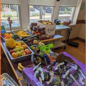 A table filled with fresh fruits and vegetables including apples, bananas, pears, carrots, strawberries, and purple-packaged snacks, set in a sunlit room with windows and a basket in the background.