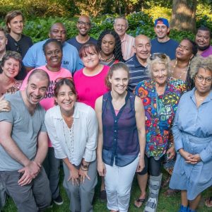 A diverse group of adults stand outdoors on grass, smiling and posing for a group photo on a sunny day, surrounded by green plants and trees.