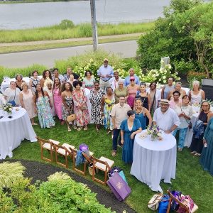 A group of people stand outdoors on a grassy lawn, smiling and facing the camera during a gathering. Cocktail tables, chairs, and bags are set up, with a lake and greenery visible in the background.