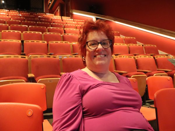 A person with short red hair and glasses, wearing a bright pink top, smiles while sitting in an empty theater with red seats.