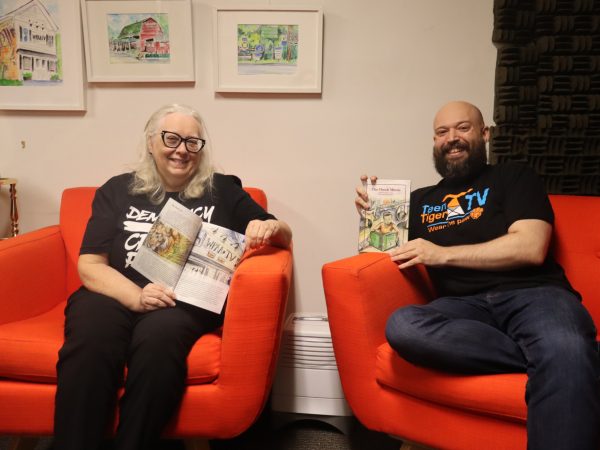 Two people sit smiling in orange chairs, each holding open illustrated books. Framed watercolor paintings hang on the wall behind them, and a small globe is visible to the left.