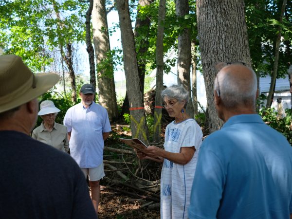 A group of older adults stands in a wooded area. A woman in a white dress is reading from a booklet while others in hats and casual clothes listen attentively in a semicircle. Sunlight filters through the trees.