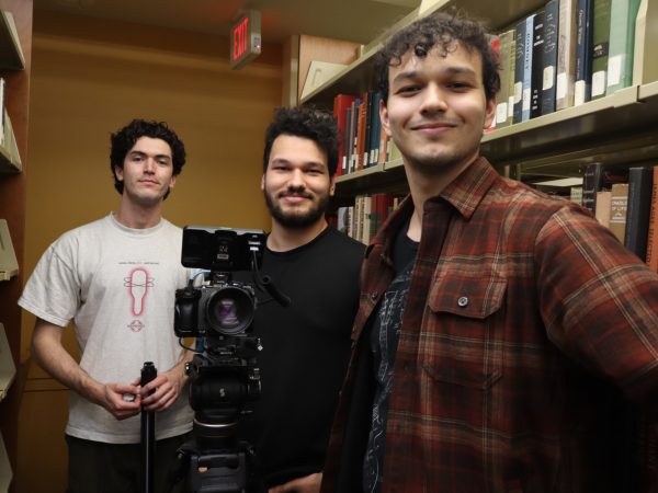 Three young men stand between bookshelves in a library, smiling at the camera. A professional video camera on a tripod is set up in front of them, suggesting they are filming or working on a media project.