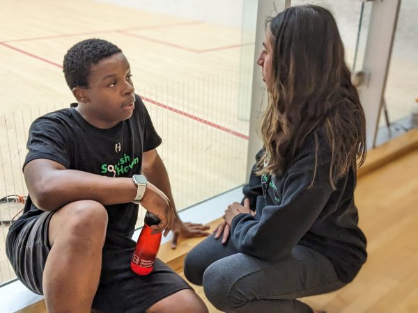 A young man and a woman sit on the floor by a glass wall of a squash court, talking. The man holds a red water bottle and wears a black Squash Haven shirt; both seem engaged in conversation.