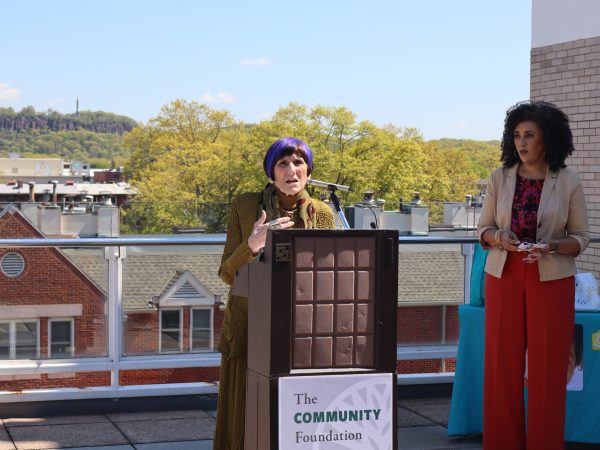 A woman with short purple hair speaks at a podium labeled The Community Foundation on a rooftop, while another woman in a beige jacket and red pants stands nearby. Trees and buildings are visible in the background.