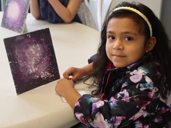 A young girl wearing a floral jacket and yellow headband sits at a table, smiling, with a painting of a purple galaxy propped up in front of her. Another child with a similar artwork is visible in the background.