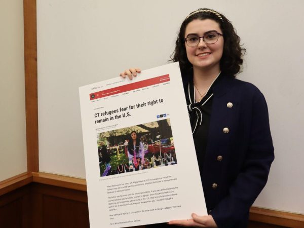 A woman with glasses and dark hair smiles while holding a large poster featuring a news article titled CT refugees fear for their right to remain in the U.S. with a photo and text.