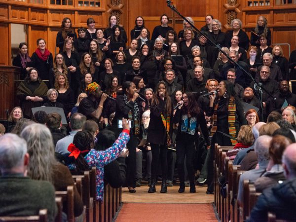 A large choir performs on stage in a wood-paneled church, with five singers in front and an audience seated in pews watching and taking photos.