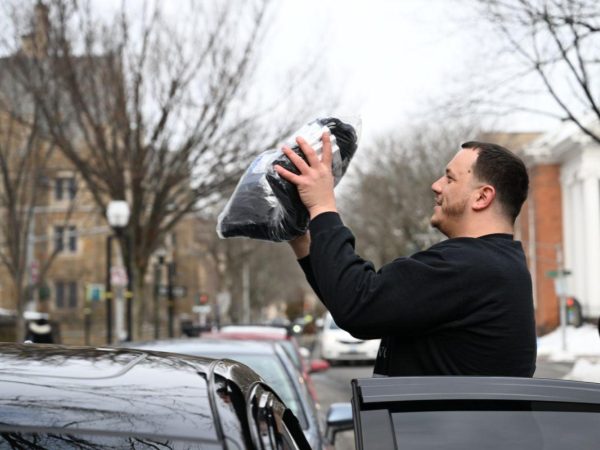 A man stands beside a car holding up a black bag with both hands on a snowy street lined with bare trees and historic buildings.