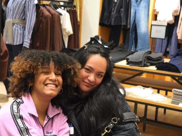 Two women smiling and posing together in a clothing store, with folded sweaters, shirts, and jackets displayed on shelves and racks behind them.