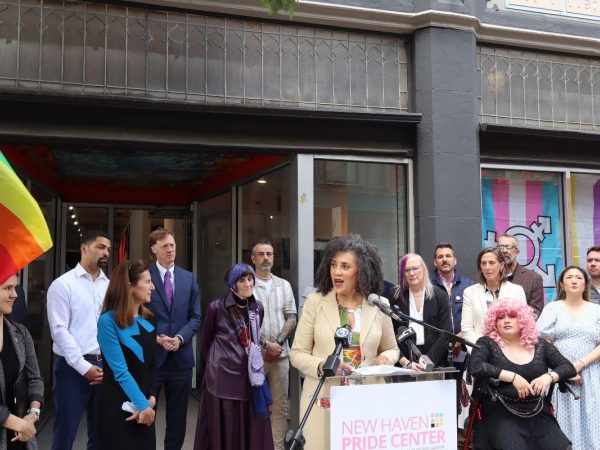 A group of people stand outside the New Haven Pride Center. A speaker addresses the crowd at a podium, with pride flags, microphones, and others listening and standing nearby. The building displays LGBTQ+ symbols and signage.