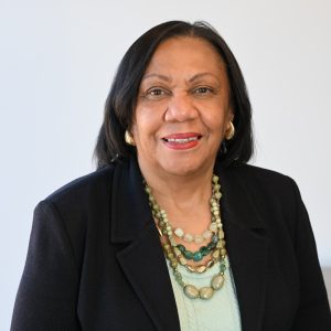 A woman with medium-length dark hair is smiling at the camera. She wears a dark blazer, a light mint top, layered green beaded necklaces, and gold hoop earrings, against a plain light background.